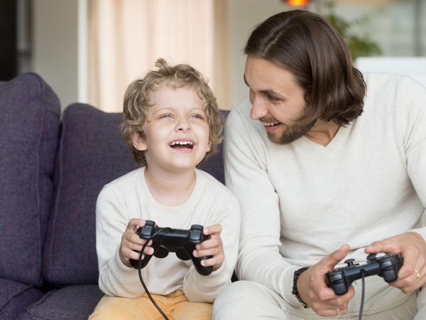 A father and his young son laugh as they play a video game together on the sofa
