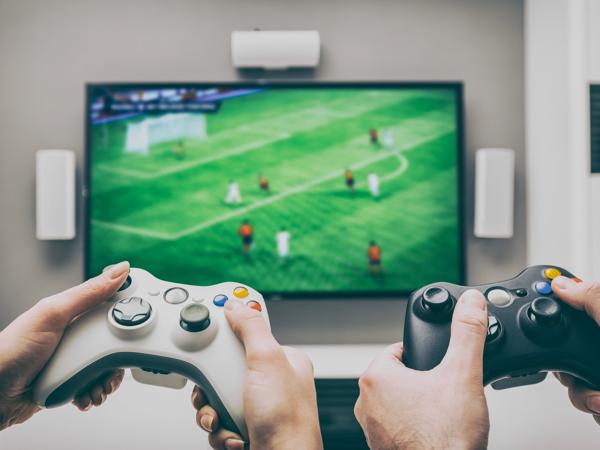 Two players hold a white and a black Xbox-style controller in front of a soccer game on TV